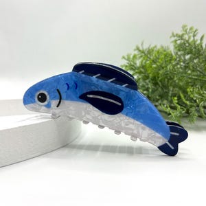 Pode incluir: Uma presilha de cabelo azul e branca em forma de peixe. A presilha apresenta um corpo azul texturizado e semitransparente com detalhes em azul escuro e uma base branca perolada. O peixe tem um olho preto e uma barbatana azul escura.