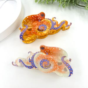 Puede incluir: Dos pinzas para el pelo con forma de pulpo. Las pinzas son de color naranja con lunares blancos y tent&aacute;culos azules. Est&aacute;n hechas de un material transl&uacute;cido. Sobre una superficie blanca.