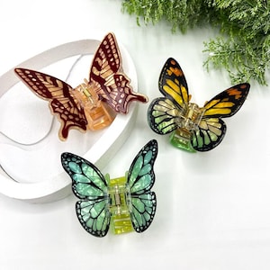May include: Three butterfly-shaped hair clips in shades of red, yellow, and teal. Each clip features intricate wing patterns and a gold-colored clasp. The clips are displayed on a white surface, with greenery in the background.