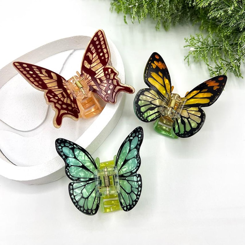 May include: Three butterfly-shaped hair clips in shades of red, yellow, and teal. Each clip features intricate wing patterns and a gold-colored clasp. The clips are displayed on a white surface, with greenery in the background.