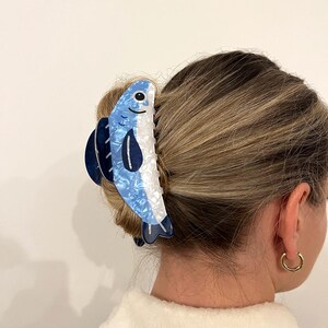 Pode incluir: Uma presilha de cabelo em forma de peixe azul e branco. A presilha apresenta um design detalhado com um rosto sorridente e detalhes em azul escuro. O acess&oacute;rio de cabelo &eacute; feito de um material brilhante e estampado.