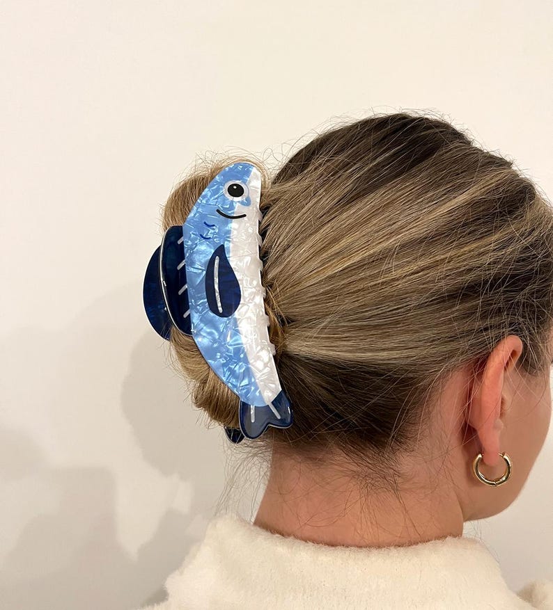 Pode incluir: Uma presilha de cabelo em forma de peixe azul e branco. A presilha apresenta um design detalhado com um rosto sorridente e detalhes em azul escuro. O acess&oacute;rio de cabelo &eacute; feito de um material brilhante e estampado.