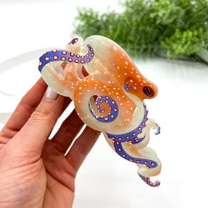 Puede incluir: Un colgante de pulpo transl&uacute;cido con manchas naranjas y blancas, detalles de tent&aacute;culos azules y blancos y un ojo rojo. El pulpo se sostiene sobre un fondo blanco, mostrando su intrincado dise&ntilde;o y colores vibrantes.