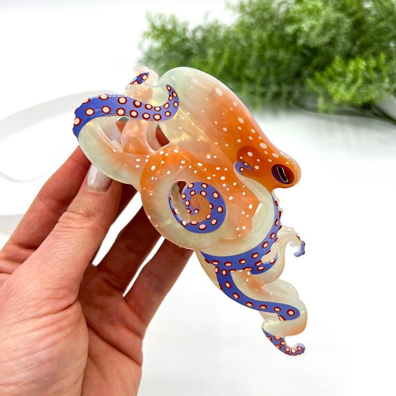 Puede incluir: Un colgante de pulpo transl&uacute;cido con manchas naranjas y blancas, detalles de tent&aacute;culos azules y blancos y un ojo rojo. El pulpo se sostiene sobre un fondo blanco, mostrando su intrincado dise&ntilde;o y colores vibrantes.