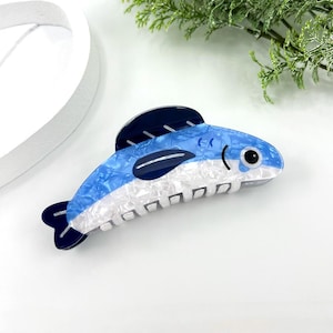 Pode incluir: Uma presilha de cabelo em forma de peixe azul e branco. A presilha apresenta um design marmorizado azul e branco no corpo, com barbatanas azuis escuras e uma base branca. &Eacute; projetada para prender o cabelo.