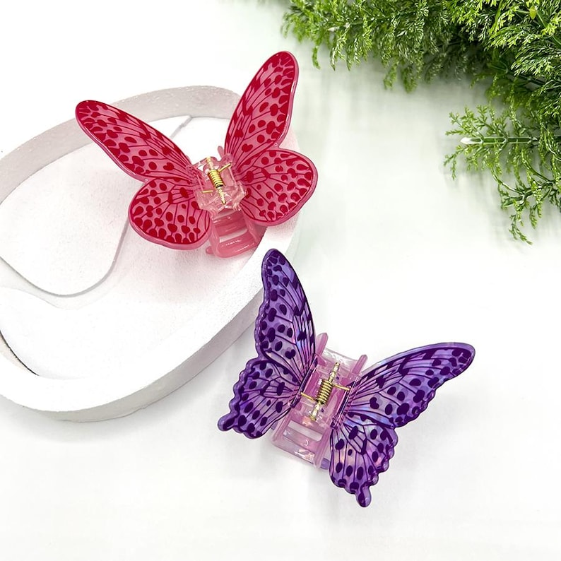 May include: Two butterfly hair clips. One is pink with darker pink spots, the other is purple with darker purple spots. Both have a clear plastic base and gold-colored metal accents. The clips are on a white surface.