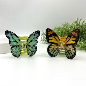 May include: Two butterfly-shaped hair clips. One clip features green and black wings, the other has yellow, orange, and black wings. Both have a gold-colored clasp and are set against a white background with greenery.