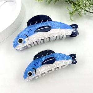 Pode incluir: Duas presilhas de cabelo em forma de peixe azul e branco. As presilhas t&ecirc;m uma barbatana e cauda azul escuro, com um corpo branco e azul. As presilhas s&atilde;o feitas de um material brilhante e estampado. As presilhas s&atilde;o projetadas para segurar o cabelo no lugar.