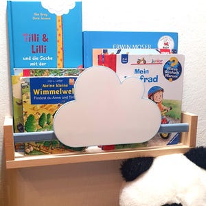 Könnte beinhalten: Ein Holzregal mit Wolken-Dekoration, das mehrere Kinderbücher enthält. Die Bücher haben blaue, gelbe und grüne Einbände mit deutschem Text. Ein Stoffpanda ist teilweise unter dem Regal zu sehen.