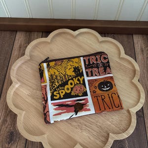 May include: A rectangular fabric zipper pouch featuring a multi-panel Halloween print with orange, yellow, and black colors. The design includes a scarecrow, a jack-o'-lantern, and text that reads "to be SPOOKY" and "TRICK OR TREAT".