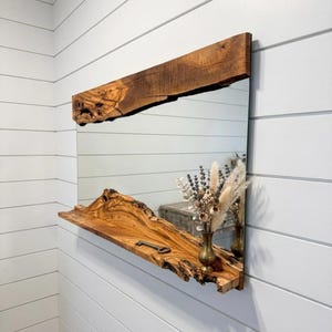May include: A rectangular mirror with a live edge wooden frame and shelf. The wood has a natural, warm brown colour. A small brass vase with dried flowers and a decorative key are on the shelf. The mirror is mounted on a white wall.