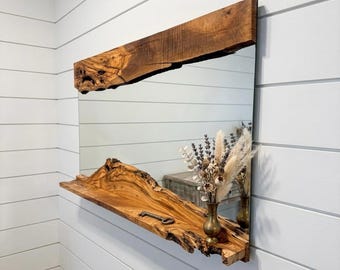 May include: A rectangular mirror with a live edge wooden frame and shelf. The wood has a natural, warm brown color. A small brass vase with dried flowers and a decorative key are on the shelf. The mirror is mounted on a white wall.