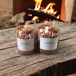 May include: Two glass jar candles with a s'mores theme. Each candle has a brown wax base, topped with whipped cream, graham crackers, marshmallows, and chocolate pieces. The labels read "SMORES" and "Organic Coconut/Soy Wax".