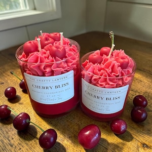 May include: Two glass candles filled with pink wax piped to resemble frosting, topped with red cherry shapes. The labels read The Crafty Lantern, Cherry Bliss, Organic Coconut/Soy Wax, 17 oz or 10 oz, and Hand Poured in Texas. Fresh cherries surround the candles.