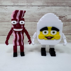 May include: Two whimsical, handmade figures: a strip of bacon with pink and red stripes and an egg with a yellow yolk. The bacon figure has a wide grin and the egg has a smiling face. Both figures have arms and legs.