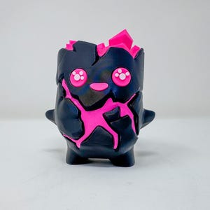 May include: A small, matte black figurine with a cracked, rock-like texture revealing vibrant neon pink material underneath. It features two round pink eyes with white accents, a small pink mouth, and jagged pink crystalline structures protruding from the top.