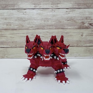 May include: A red, three-headed dog figurine with yellow eyes and white teeth. The figurine has a pixelated design and is wearing studded collars. The dog is standing on a white surface.