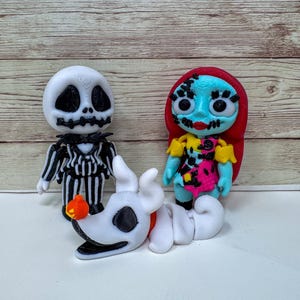 May include: A trio of figurines featuring characters from The Nightmare Before Christmas. Jack Skellington in a black and white pinstripe suit, Sally in a patchwork dress, and Zero the ghost dog. The figures are set against a light wood grain background.