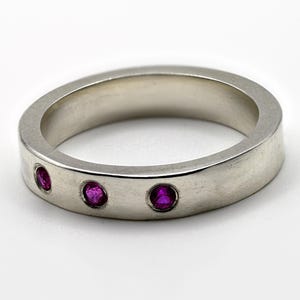 以下が含まれることがあります： 滑らかでつや消し仕上げのシルバー調の金属製バンドリング。リングの外側表面には、3つの小さな丸い鮮やかなピンク色の宝石が埋め込まれており、前面に沿って水平に一列に並んでいます。