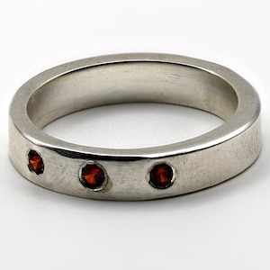 Peut inclure: Une bague anneau de couleur argentée avec une finition lisse et polie, ornée de trois petites pierres précieuses rondes d'un rouge profond incrustées à fleur de métal. L'anneau présente une largeur uniforme et une forme circulaire.