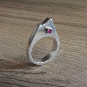 Könnte beinhalten: Ein silberfarbener Ring mit glatter, gebürsteter Metalloberfläche und einem spitz zulaufenden, architektonischen Oberteil. Ein kleiner, runder, tiefrosa Edelstein ist in den oberen Teil der Ringschiene eingefasst und bildet einen Blickfang.