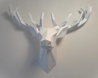Könnte beinhalten: Eine weiße, geometrische Hirschkopf-Wanddekoration. Die Skulptur hat ein polygonales Design mit scharfen Winkeln und eine symmetrische Form. Das Geweih erstreckt sich nach außen, und der Kopf ist an einer neutralfarbenen Wand befestigt.