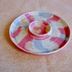 May include: A ceramic serving dish with a central dip bowl. The dish is painted with a colorful pattern of pink, white, yellow, and blue brushstrokes. The dish is round and has a glossy finish.