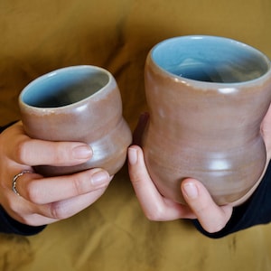 Puede incluir: Dos tazas de cerámica hechas a mano. El exterior es de color marrón cálido con un acabado brillante, mientras que el interior es azul claro. Las tazas tienen una forma curva única y las sostiene una persona.