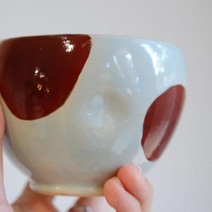 May include: A small, rounded ceramic bowl with a glossy, light blue base glaze. The exterior features large, irregular, dark reddish-brown spots painted onto the surface. The bowl has a smooth, curved shape with a slightly indented texture.