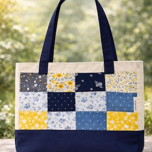 May include: A rectangular tote bag with navy blue handles and base. The upper section features a patchwork design with squares of floral and dotted patterns in yellow, blue, and gray. The bag is made of fabric.