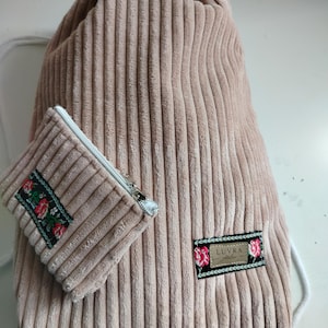 Könnte beinhalten: Ein Kordelzug-Rucksack und eine passende Reißverschlusstasche aus rosa Cord. Beide Artikel haben eine gerippte Textur und floral bestickte Aufnäher. Die Tasche hat einen silbernen Reißverschluss. Der Rucksack hat einen weißen Kordelzug.