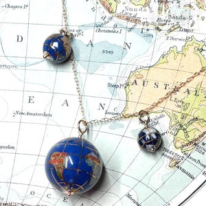 May include: A gold-coloured necklace with three globe pendants. The blue globes with gold continents hang from fine chains. The necklace is set against a vintage map, evoking a sense of travel and exploration.