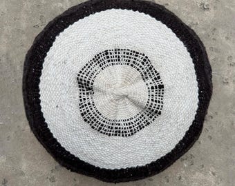 May include: Circular woven rug with a black border, a white center, and a black patterned design in the middle. The rug is made of natural fibers and has a textured appearance. The rug is suitable for indoor use.