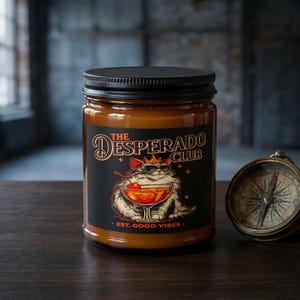 Op de afbeelding: Een kaars in een glazen pot met een zwart deksel. Het etiket toont een kat met een zonnebril en een kroon, die een cocktail vasthoudt. Tekst op het etiket luidt "The Desperado Club" en "Est. Good Vibes."