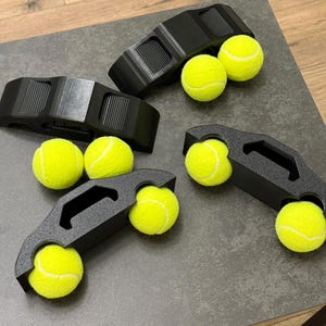 以下が含まれることがあります： 4つの黒いテニスボールホルダー。それぞれに2つの黄色のテニスボールが入っています。ホルダーはマットブラック素材でできており、ハンドル付きの湾曲したデザインです。テニスボールは明るい黄色でふわふわしています。