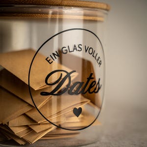 Könnte beinhalten: Ein klares Glas mit einem Holzdeckel, gefüllt mit kleinen braunen Papierumschlägen. Das Glas hat ein schwarzes rundes Etikett mit den Worten "EIN GLAS VOLLER Dates" und einem schwarzen Herzsymbol.
