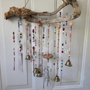 Puede incluir: Un móvil de viento artesanal con una rama de madera flotante natural, adornado con hilos de cuentas coloridas, pequeñas campanas y carillones de metal. Las cuentas son de varios colores y formas, creando una estética bohemia.