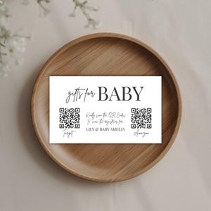 Op de afbeelding: Een witte kaart met de tekst "gifts for BABY" in een scriptlettertype. Hieronder staan twee QR-codes met de woorden "Target" en "Amazon". De kaart bevat ook de tekst "Kindly scan the QR Codes to view the registries for LILY & BABY AMELIA."
