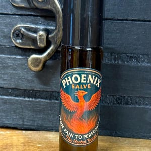 May include: A small, dark glass bottle with a black cap. The label reads "Phoenix Salve" with a graphic of a red phoenix. The text "From Pain to Perform" and "The Herbal Hedge" are also on the label. The bottle is on a wooden surface.