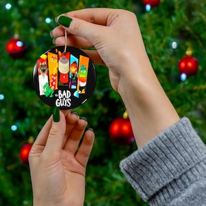 May include: A round, black Christmas ornament featuring characters from "The Bad Guys" movie. The ornament has a gold string for hanging and is held against a backdrop of a Christmas tree with red ornaments.