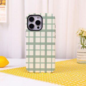May include: A smartphone case featuring a light cream background with a sage green grid pattern. The case has a rectangular cutout at the top left corner for a triple-lens camera system, with a black border surrounding the camera lens area.