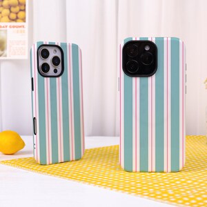 May include: Two smartphone cases featuring a vertical striped pattern in alternating shades of teal, white, and thin pink lines. Each case has a black camera cutout area with three circular lenses and a flash, designed to fit a modern mobile device.