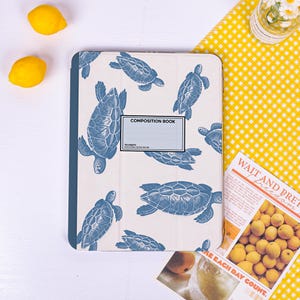 May include: A tablet case featuring a cream-coloured background with a repeating pattern of blue sea turtles. A central rectangular label reads COMPOSITION BOOK, 100 SHEETS, 9.75 X 7.5 IN / 24.76 X 19.05 CM. The case has a solid dark blue spine.