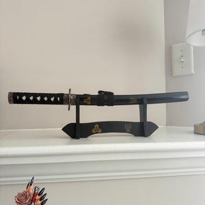 May include: A black katana sword with a decorative handle and a black scabbard, displayed on a black stand. The handle is wrapped with a black cord. The stand has gold-colored accents. The sword is displayed on a white shelf.