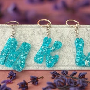 Può includere: Tre portachiavi in resina color acquamarina che compongono le lettere "MKL". Ogni lettera è riempita di scaglie d'argento e attaccata a un portachiavi d'argento. I portachiavi sono esposti su una superficie grigio chiaro con fiori viola in primo piano.