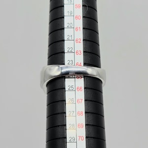 May include: A silver ring is placed on a black ring sizer. The ring is a simple band design. The ring sizer has measurements in both inches and millimeters. The ring is being measured for size.