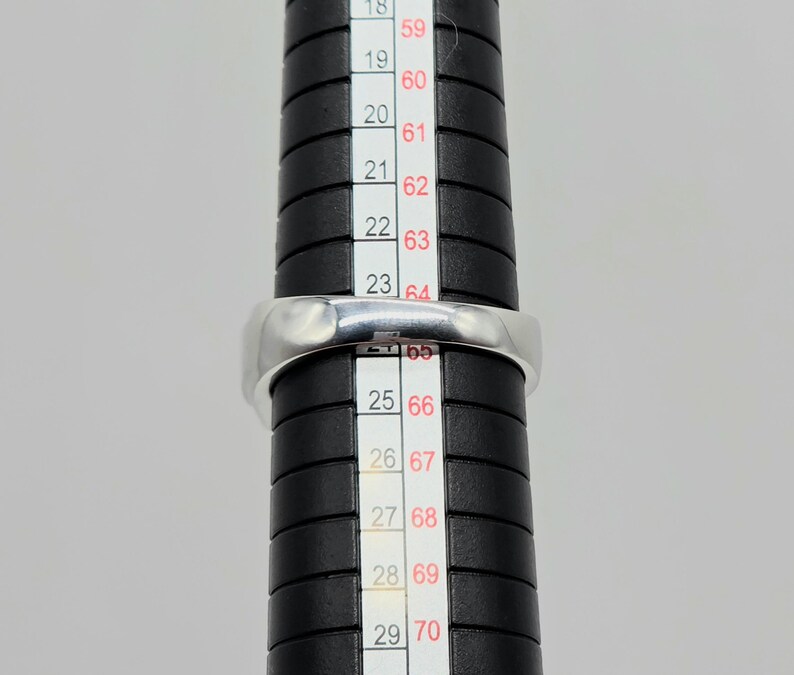 May include: A silver ring is placed on a black ring sizer. The ring is a simple band design. The ring sizer has measurements in both inches and millimeters. The ring is being measured for size.