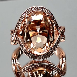May include: A rose gold ring featuring a large, oval-cut, champagne-colored center stone. The setting is surrounded by a halo of small, clear, pavé-set stones, with additional small stones embedded along the twisted, crossover-style band.