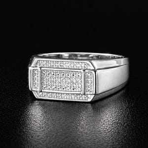 May include: A silver ring with a rectangular face, encrusted with small, sparkling stones. The ring has a polished finish and a classic, masculine design, suitable for everyday wear or special occasions.