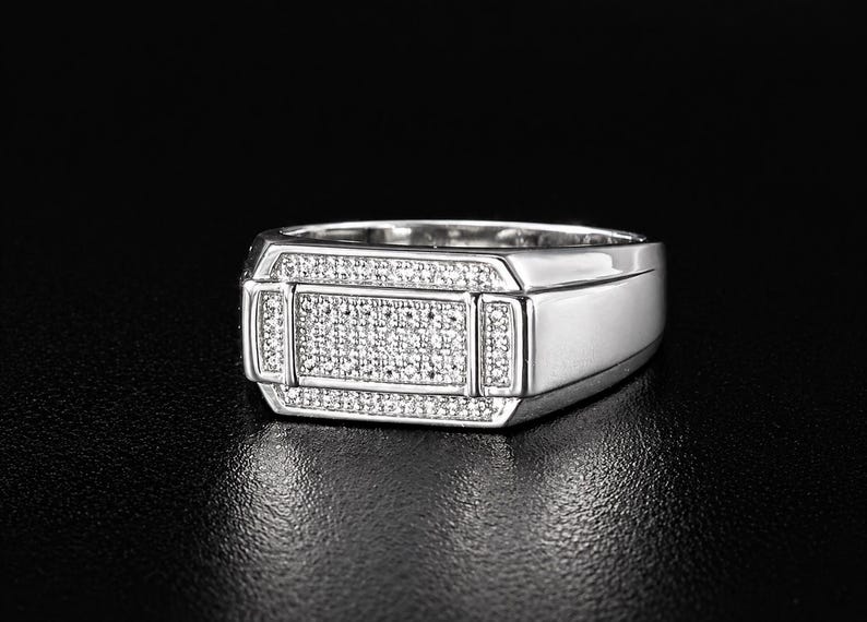 May include: A silver ring with a rectangular face, encrusted with small, sparkling stones. The ring has a polished finish and a classic, masculine design, suitable for everyday wear or special occasions.